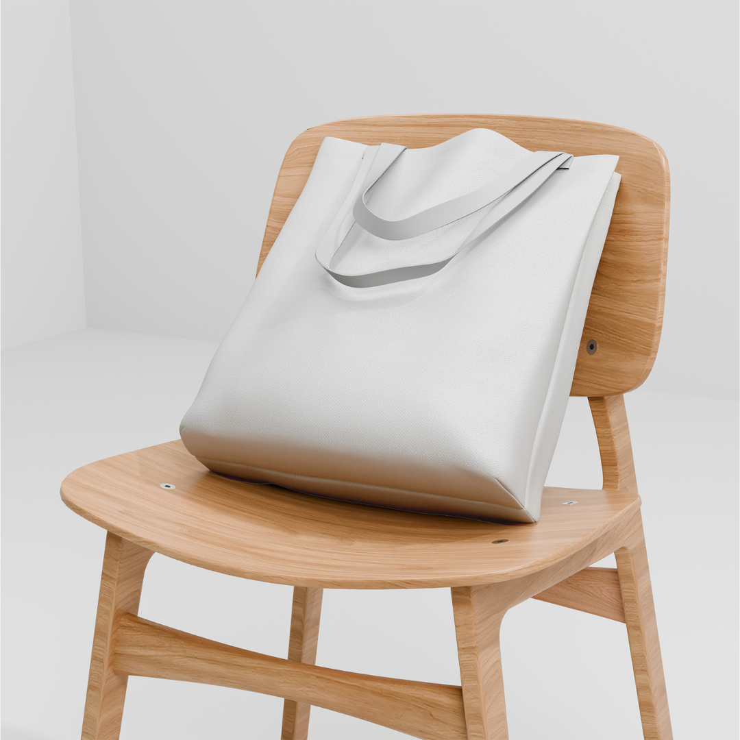 White canvas tote bag with shoulder straps leaning against the back of a minimalist light wood chair against a neutral grey background